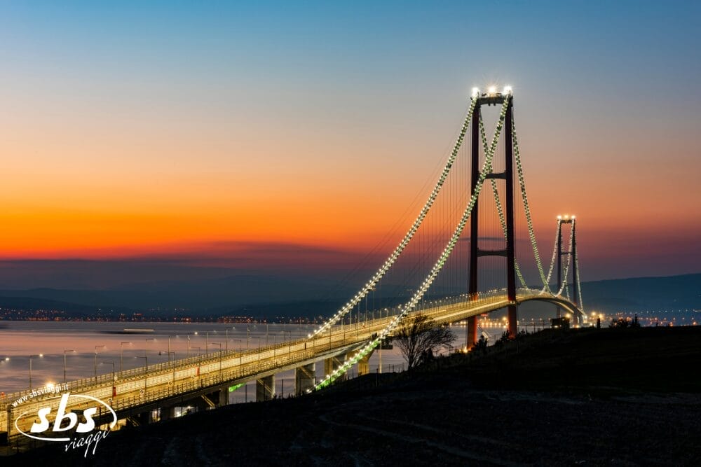 Un ponte sospeso illuminato si estende su acque tranquille al tramonto, sotto un cielo blu e arancione. Le luci della città scintillano in lontananza e nell'angolo in basso a sinistra compare il logo "sbs" con il marchio automatico Bozza.