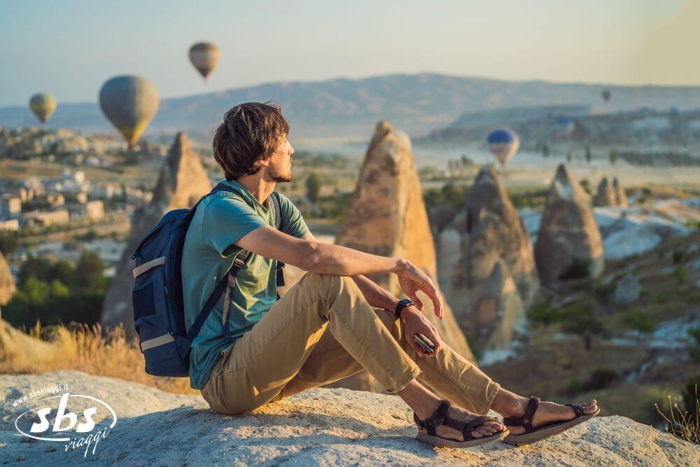 Un uomo con lo zaino in spalla siede su una roccia e osserva le mongolfiere che fluttuano su un paesaggio panoramico con formazioni rocciose uniche e montagne lontane, catturando la magia della bozza automatica durante il giorno.