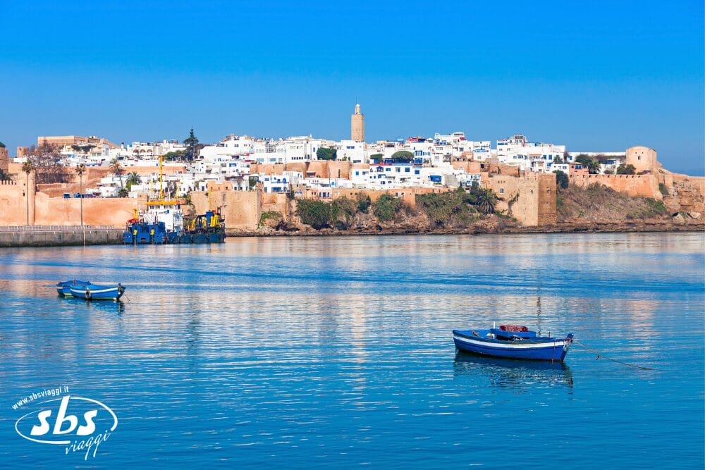 Acqua azzurra e calma con due barche, un muro fortificato e una città di edifici bianchi e un'alta torre sullo sfondo sotto un cielo azzurro e limpido: scoprite il fascino delle Città Imperiali del Marocco nella vostra prossima avventura Fly & Drive.