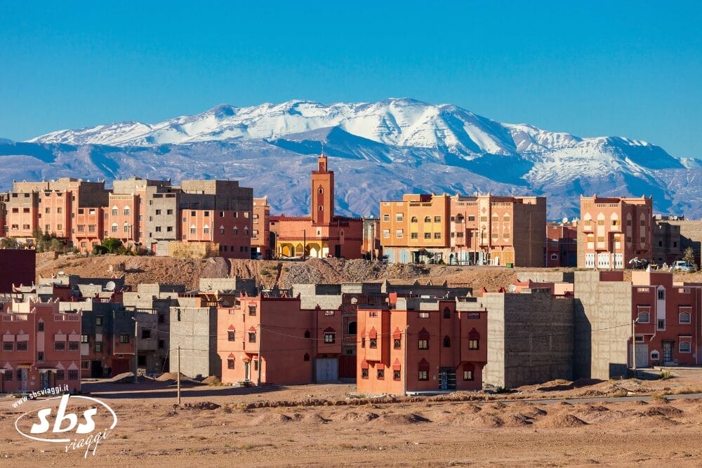 Una città con edifici in terracotta e beige si trova di fronte a montagne innevate sotto un cielo azzurro e limpido. Tra gli edifici si erge un'alta torre dell'orologio, mentre il primo piano mostra un terreno secco e sabbioso: una classica scena di Bozza automatica.