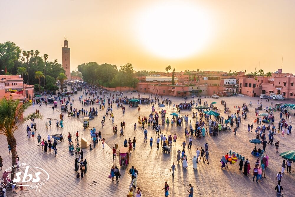 Una grande e animata piazza di Marrakech al tramonto, piena di gente che passeggia e si riunisce. L'area è fiancheggiata da edifici rosa e alberi verdi, con un'alta torre della moschea sullo sfondo, che cattura il cuore vibrante del Marocco a pochi passi da un riad tradizionale.