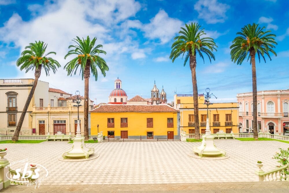 Una piazza soleggiata con alte palme, edifici storici colorati e cupole di chiese sullo sfondo, sotto un cielo azzurro con nuvole sparse, crea una scena invitante che ricorda una cartolina di Bozza automatica.