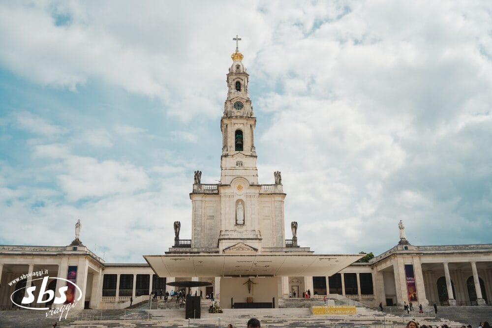 L'immagine mostra il Santuario di Fátima, in Portogallo, con la sua alta basilica centrale con torre a crociera, ingresso ad arco e colonnati, sotto un cielo parzialmente nuvoloso, ripreso come Bozza automatica.