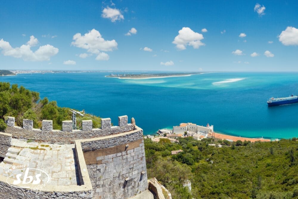 Vista panoramica da una fortezza in pietra che domina una città costiera, con mare turchese, banchi di sabbia, colline verdi e una nave attraccata: una scena idilliaca sotto un cielo blu brillante con nuvole sparse. Bozza automatica.