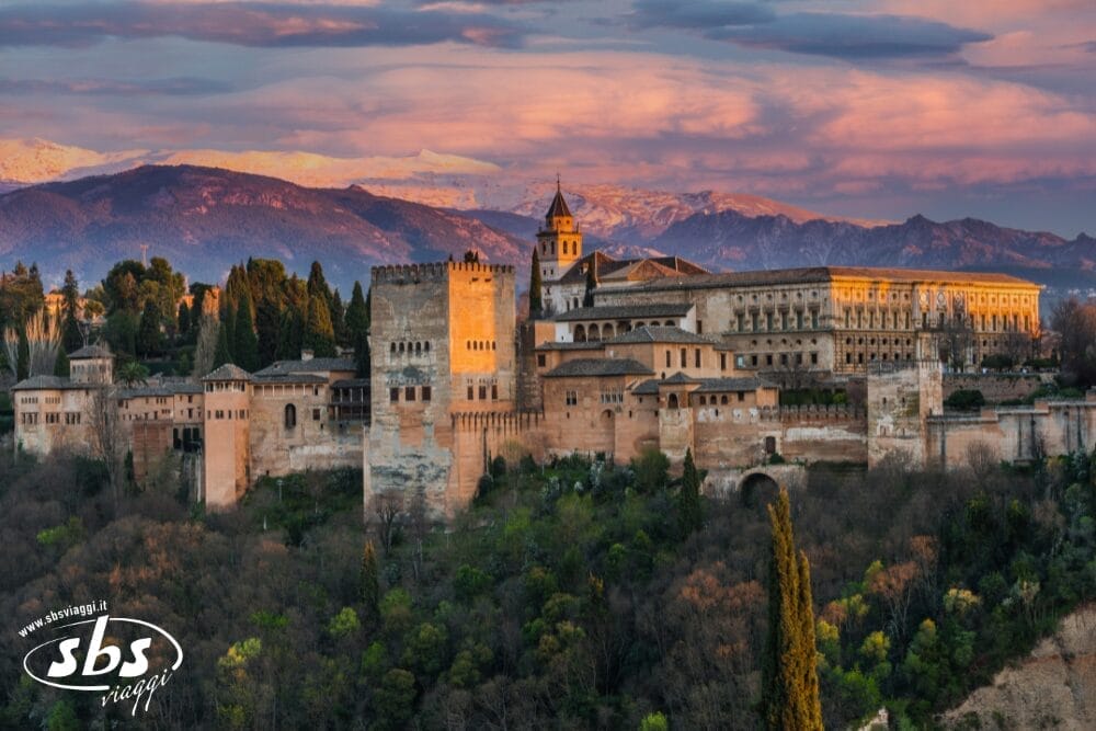 Il palazzo e il complesso della fortezza dell'Alhambra a Granada, in Spagna, con le mura e le torri illuminate dal tramonto, circondate da una vegetazione lussureggiante e dalle montagne sullo sfondo, sotto un cielo colorato: un'attrazione iconica di qualsiasi Gran Tour dell'Andalusia.