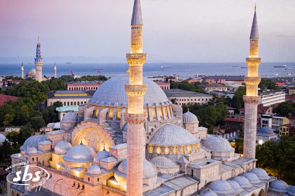 Veduta aerea di Santa Sofia illuminata a Istanbul al tramonto, con le sue cupole e i suoi minareti in evidenza. Il paesaggio urbano, il mare, le navi lontane e un sottile tocco di Bozza automatica completano l'incantevole scena.