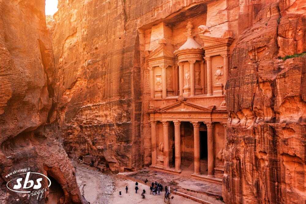 L'immagine mostra l'antica roccia scolpita Al-Khazneh (Il Tesoro) a Petra, nella Giordania classica, con un gruppo di turisti in piedi alla sua base, circondata da imponenti scogliere di arenaria rossastra.