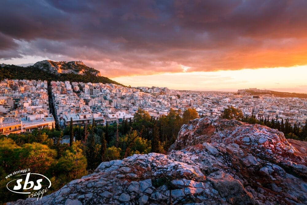 Vista del tramonto su Atene, in Grecia, con nuvole drammatiche, edifici della città, primo piano roccioso, alberi verdi e colline sullo sfondo. Il cielo si illumina di sfumature arancioni e viola sopra la città: una vera Bozza automatica della bellezza della natura.