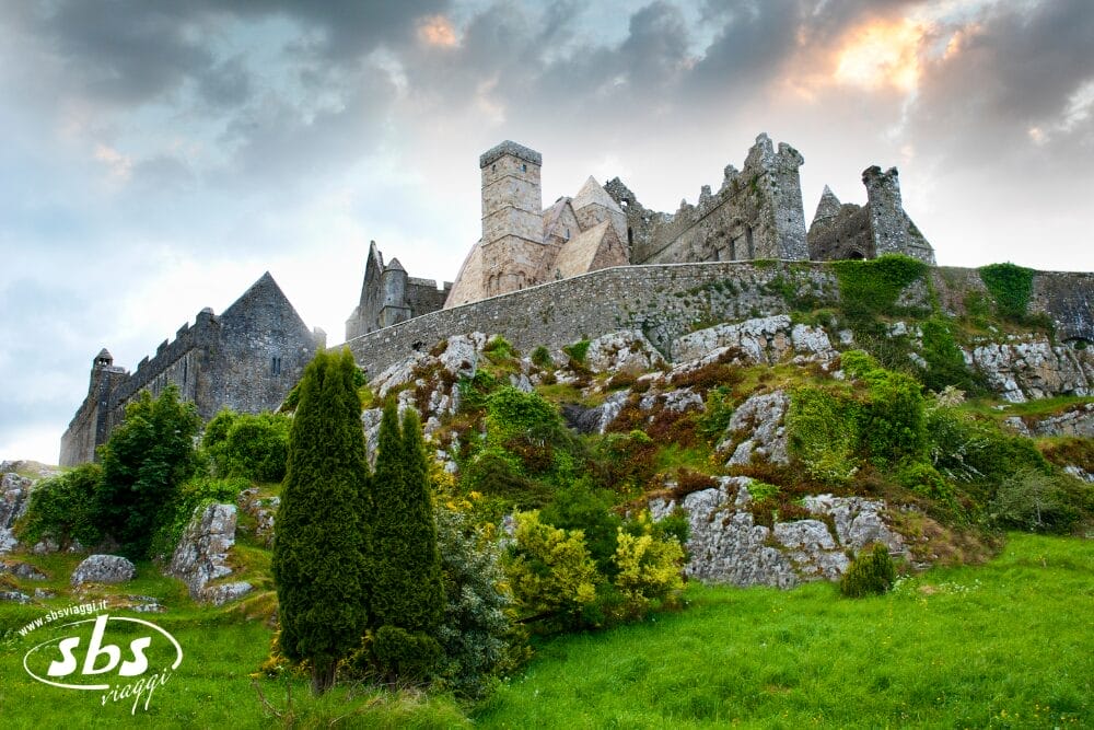 Un antico castello in pietra si trova in cima a una collina rocciosa circondata da vegetazione e arbusti, sotto un drammatico cielo nuvoloso al tramonto, che evoca lo spirito dell'Irlanda express. Il logo con "sbs" appare nell'angolo in basso a sinistra.