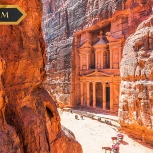 Una veduta dell'antica roccia Al-Khazneh (Il Tesoro) a Petra, in Giordania, circondata da alte scogliere rosse, con la luce del sole che evidenzia l'intricata facciata e i cammelli che riposano sotto. Nell'immagine compaiono "Bozza automatica", "PREMIUM" e il logo di SBS.