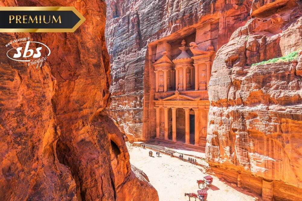 Una veduta dell'antica roccia Al-Khazneh (Il Tesoro) a Petra, in Giordania, circondata da alte scogliere rosse, con la luce del sole che evidenzia l'intricata facciata e i cammelli che riposano sotto. Nell'immagine compaiono "Bozza automatica", "PREMIUM" e il logo di SBS.