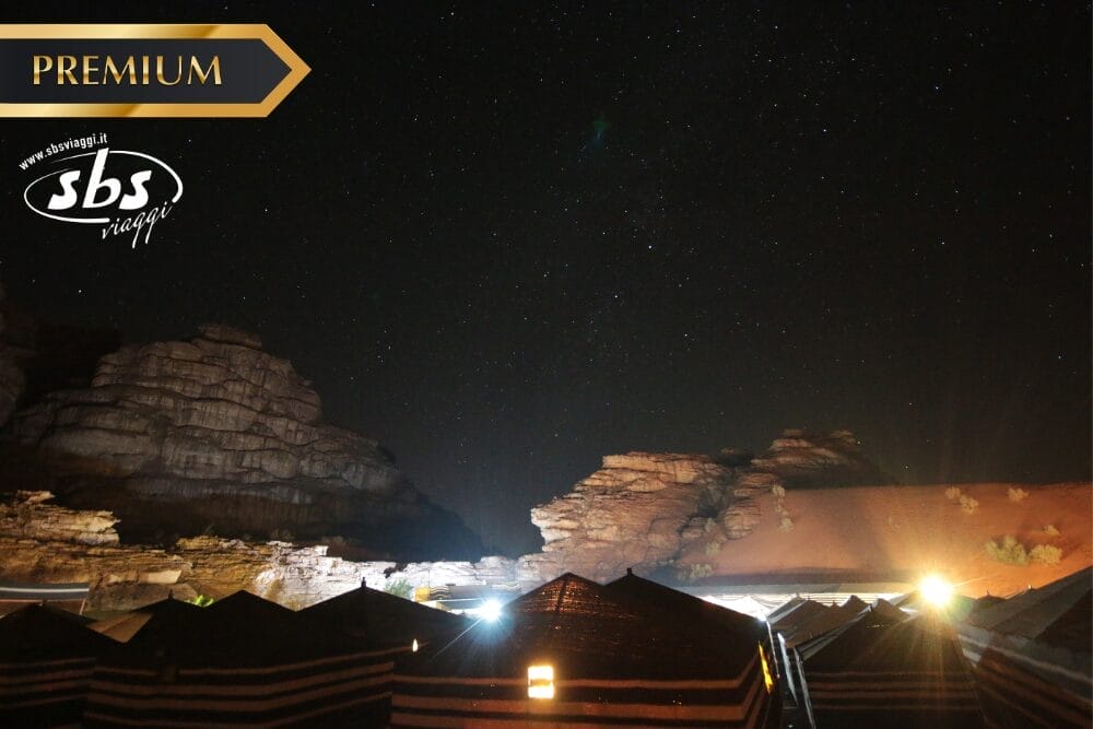Veduta notturna di scogliere rocciose del deserto sotto un cielo stellato, con tetti di tende e luci soffuse in primo piano. Nell'angolo superiore sinistro compaiono le diciture "PREMIUM", "sbs viaggi" e una sottile filigrana Bozza automatica.