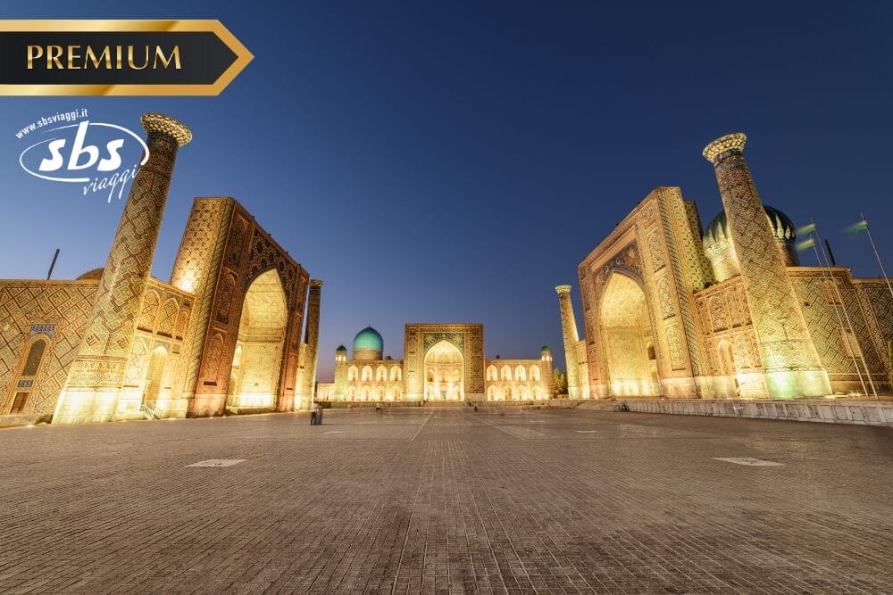 Veduta notturna di Piazza Registan a Samarcanda, Uzbekistan, con l'architettura storica islamica e le cupole illuminate; i loghi "PREMIUM", "sbs viaggi" e "Bozza automatica" appaiono nell'angolo in alto a sinistra.
