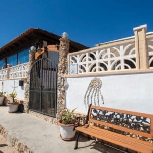 Una casa in pietra e stucco con un ingresso recintato, un muro decorativo, piante in vaso e una panchina di legno all'esterno; un mosaico di meduse decora la parete bianca sotto un cielo azzurro e limpido: una vera Bozza automatica di fascino costiero.