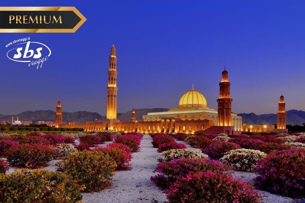 Una grande moschea illuminata con cupole e minareti si staglia in un cielo crepuscolare, circondata da vivaci giardini fioriti. Nell'angolo in alto a sinistra compaiono le scritte "PREMIUM", "sbs viaggi" e "Bozza automatica". Sullo sfondo si ergono le montagne.