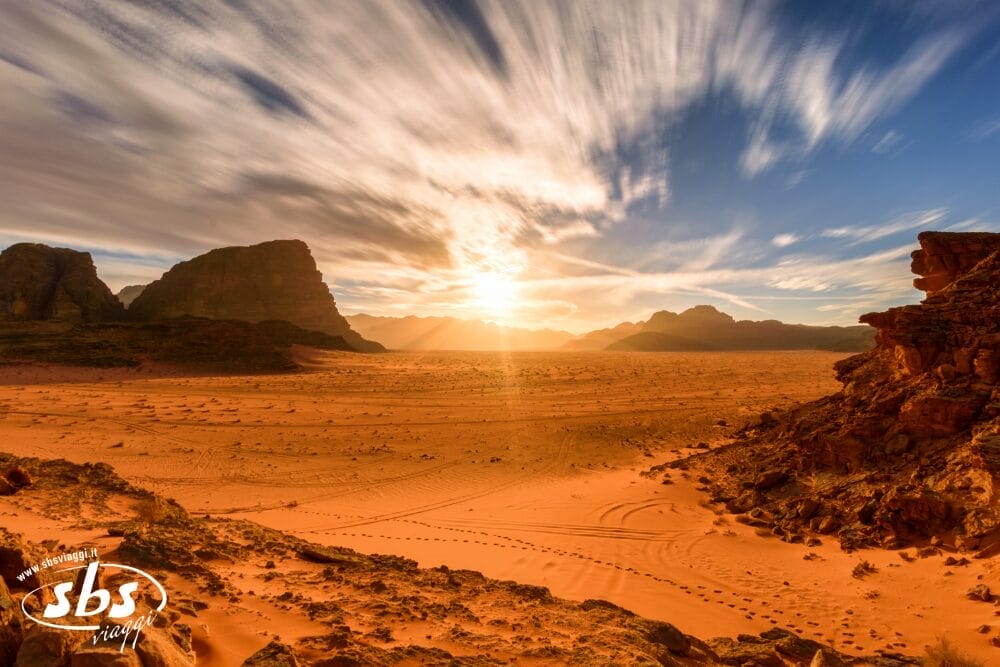 Paesaggio desertico arancione con formazioni rocciose sotto un cielo drammatico al tramonto. I raggi del sole illuminano la sabbia, rivelando le tracce di pneumatici e animali di un tour di Tende di Stelle. Il logo SBS è visibile nell'angolo in basso a sinistra.