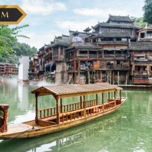 Una barca di legno galleggia su un fiume verde davanti a edifici tradizionali cinesi in legno con tetti in tegole. Il logo SBS Premium e una sottile filigrana Bozza automatica appaiono mentre il verde lussureggiante circonda la scena pacifica.