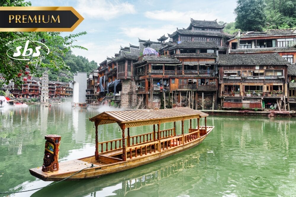 Una barca di legno galleggia su un fiume verde davanti a edifici tradizionali cinesi in legno con tetti in tegole. Il logo SBS Premium e una sottile filigrana Bozza automatica appaiono mentre il verde lussureggiante circonda la scena pacifica.