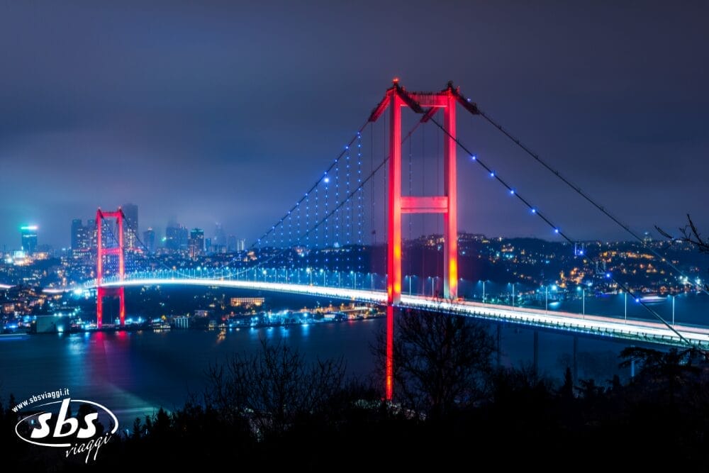 Un ponte sospeso illuminato con torri rosse e luci blu attraversa un fiume di notte, con lo skyline della città sullo sfondo. La scena calma e nebbiosa sembra quasi surreale, come una bozza automatica di bellezza urbana.
