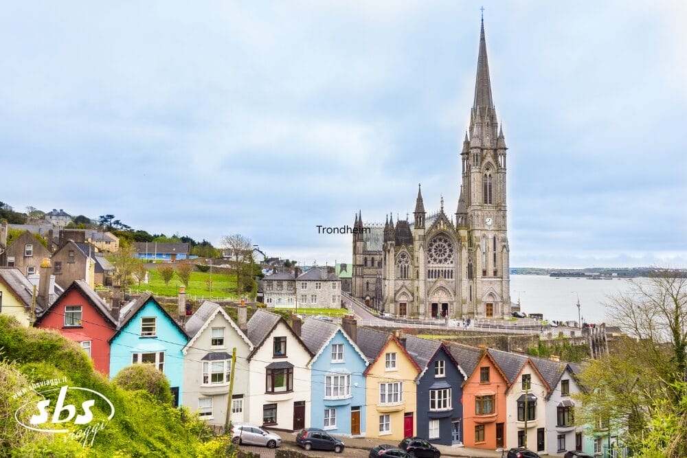 Una fila di case colorate si trova su una strada in pendenza di fronte a una grande chiesa in stile gotico con un'alta guglia, una scena perfetta da immortalare durante il vostro gran tour d'Irlanda, affacciata su acque tranquille sotto un cielo nuvoloso.