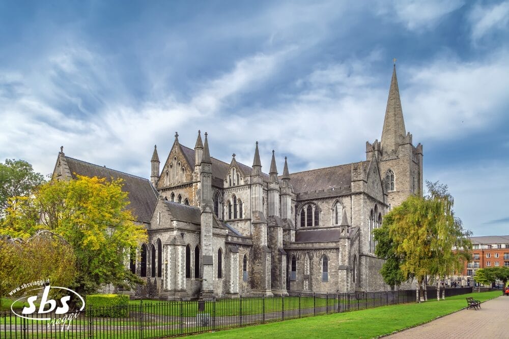 Una grande cattedrale in pietra in stile gotico con archi a sesto acuto, un'alta guglia e dettagli intricati si erge maestosa lungo il percorso dell'Irlanda Express, circondata da un prato verde, alberi e una recinzione metallica nera sotto un cielo parzialmente nuvoloso.