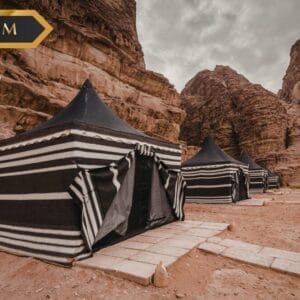 Tre tende a strisce bianche e nere si trovano in un deserto roccioso con alte scogliere sotto un cielo nuvoloso. Sull'immagine sono presenti l'etichetta "PREMIUM", il logo "sbs" e la targhetta Bozza automatica.