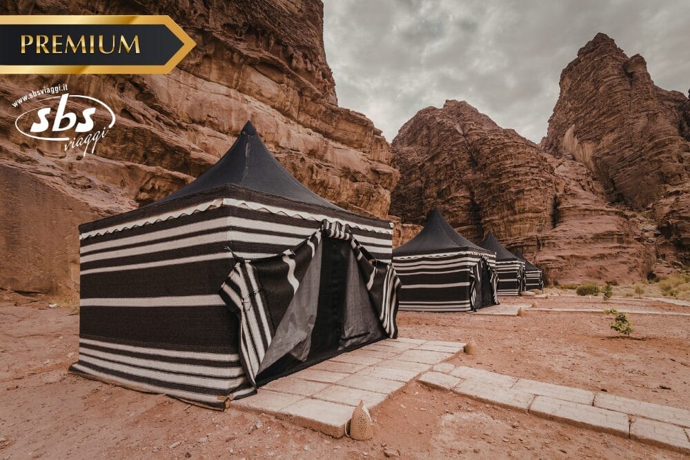 Tre tende di lusso a strisce bianche e nere sono montate su un sentiero di pietra tra le rocce del deserto sotto un cielo nuvoloso. Un banner "PREMIUM", il logo "sbs" e il testo "Bozza automatica" sono visibili negli angoli superiori dell'immagine.