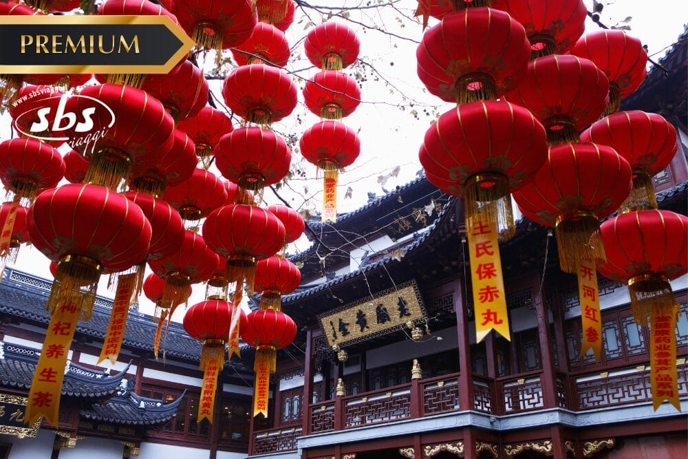 Lanterne cinesi rosse pendono sopra un tempio tradizionale con tetti ornati e insegne dorate, creando un'atmosfera di festa. I loghi "PREMIUM", "sbs" e "Bozza automatica" si trovano nell'angolo in alto a sinistra, a indicare una speciale celebrazione culturale.