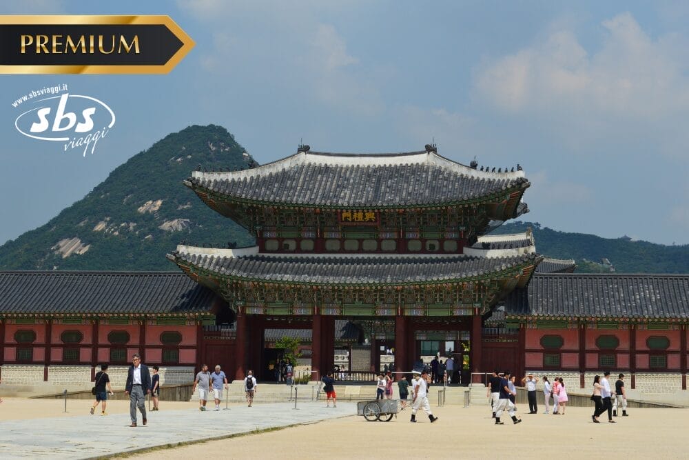 Turisti camminano davanti al cancello principale di un palazzo tradizionale coreano con un paesaggio montano sullo sfondo; i loghi "PREMIUM", "Bozza automatica" e "sbs viaggi" appaiono sull'immagine.