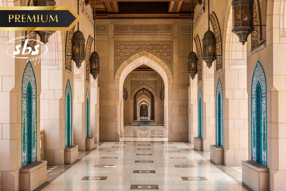 Un grande corridoio con archi ornati, piastrelle a motivi, finestre colorate di blu e lanterne pendenti, che mostrano l'architettura islamica. I loghi "PREMIUM", "SBS" e "Bozza automatica" appaiono nell'angolo superiore sinistro.