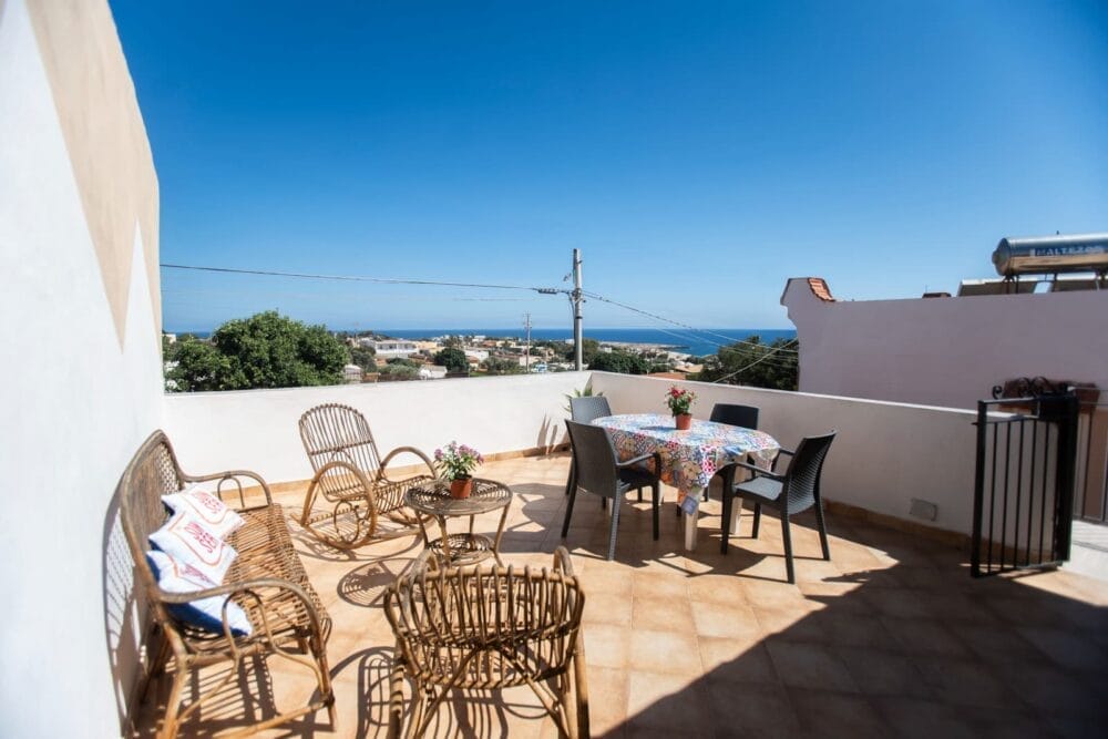 Terrazza soleggiata sul tetto con sedie in vimini, un tavolo coperto da una tovaglia floreale, piante in vaso e una splendida vista automatica Bozza sul mare e sugli edifici lontani sotto un cielo azzurro e limpido.
