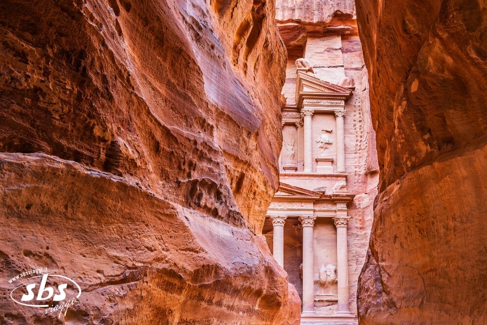 L'antica facciata in pietra del Tesoro di Petra è visibile attraverso le strette pareti di roccia rossastra del Siq, con la luce del sole che mette in risalto gli intricati intagli. Il logo "sbs" e il testo "Tour Giordania Discovery" appaiono nell'angolo in basso a sinistra.