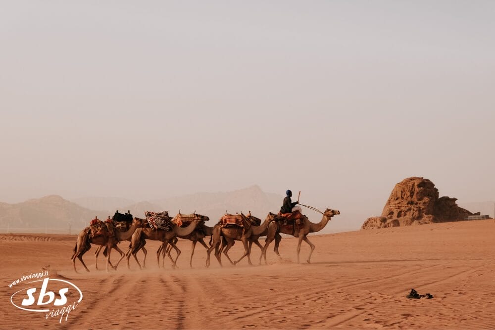 Una persona cavalca un cammello, guidando una carovana sulla sabbia rossastra sotto un cielo nebuloso, con montagne lontane e tracce di pneumatici. Il logo SBS Viaggi e "Tour Tende di Stelle" evocano l'avventura in questa scena desertica.