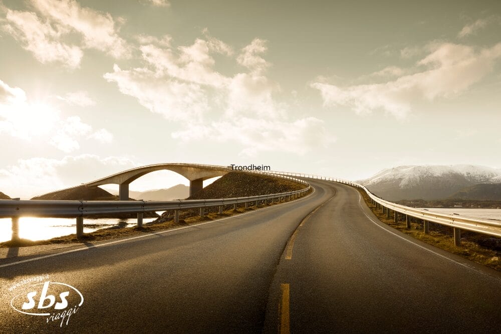 Una strada curvilinea e un drammatico ponte ad arco conducono verso montagne lontane, sotto un cielo nuvoloso, immerso in una luce morbida e dorata. Sopra il ponte appare la scritta "Trondheim", incastonata nei panoramici fiordi della Norvegia lungo la famosa Via Atlantica.