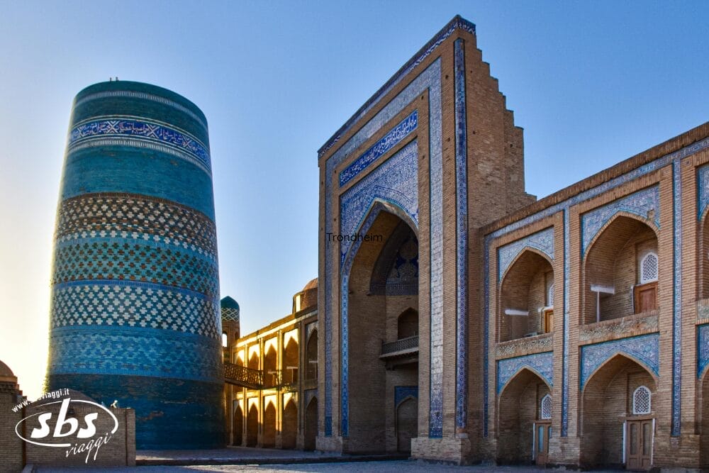 Un sito storico in Uzbekistan con un grande minareto cilindrico a piastrelle blu e un edificio adiacente in mattoni con intricate piastrelle blu e porte ad arco, immerso in una calda luce solare: un vero gioiello lungo la Via della Seta.