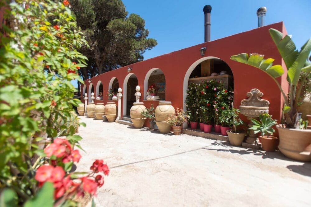 Un edificio in stile mediterraneo con pareti rosse, portali ad arco e grandi vasi d'argilla pieni di piante si trova sotto un cielo azzurro e limpido: il B&B Paradise è circondato da una vegetazione lussureggiante e da vivaci piante fiorite.