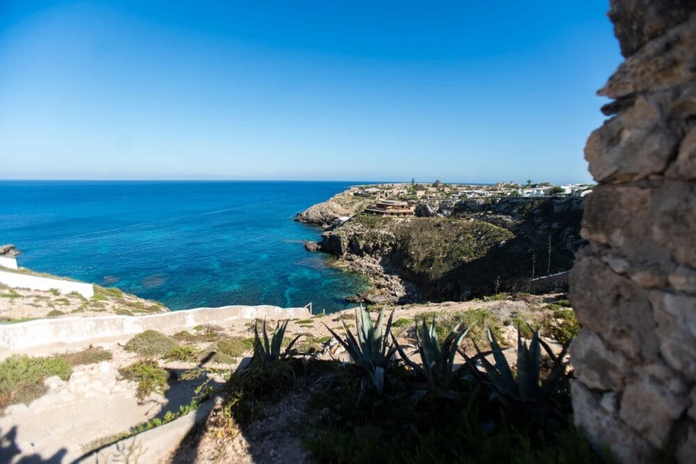 Veduta di una costa rocciosa con un mare azzurro e limpido sotto un cielo luminoso, alcune piante verdi e agave in primo piano e un muro di pietra sulla destra. In lontananza, edifici appollaiati sulle scogliere sovrastano questa pittoresca scena di Bozza automatica.