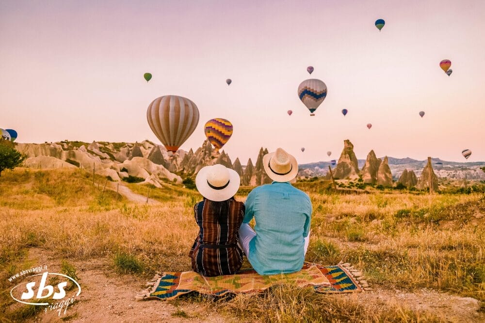 Una coppia con il cappello siede su una coperta in un campo erboso, ammirando formazioni rocciose uniche e diverse mongolfiere colorate che fluttuano nel cielo al tramonto: un perfetto momento di bozza automatica.
