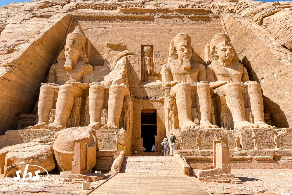 Quattro grandi statue sedute scolpite nella facciata rocciosa del tempio di Abu Simbel, in Egitto, con alcune persone in piedi all'ingresso, evidenziano l'imponenza e l'architettura antica del tempio: una vera Bozza automatica di arte monumentale.