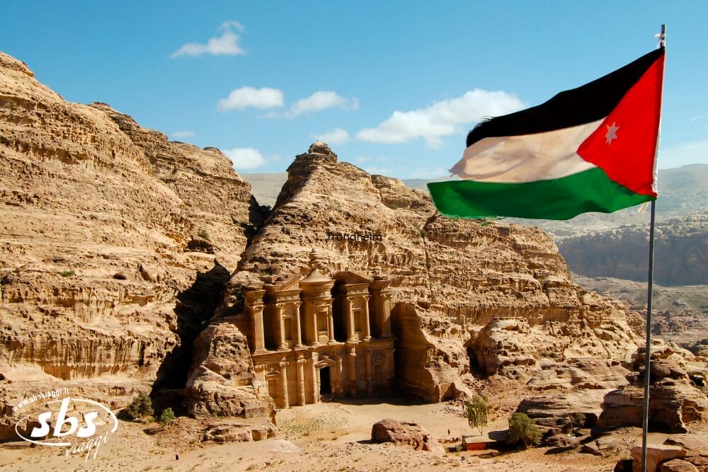 Una grande bandiera giordana sventola in primo piano, mentre l'antico monastero di Petra, scolpito nella roccia, è incastonato tra aspre scogliere sabbiose sotto un cielo blu brillante: un'attrazione straordinaria di qualsiasi tour della Giordania classica.
