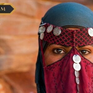 Una persona che indossa un foulard tradizionale e copre il viso con monete d'argento, con i soli occhi visibili. Lo sfondo è sfocato in toni caldi e terrosi. I loghi "PREMIUM", "sbs viaggi" e "Bozza automatica" appaiono nell'immagine.