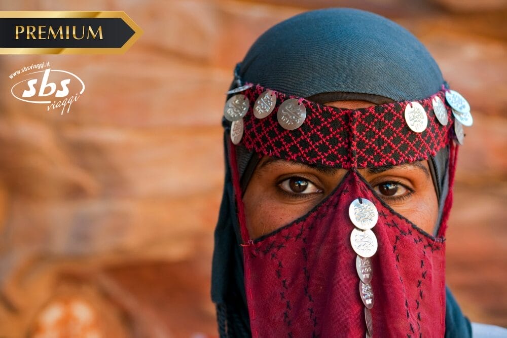 Una persona che indossa un foulard tradizionale e copre il viso con monete d'argento, con i soli occhi visibili. Lo sfondo è sfocato in toni caldi e terrosi. I loghi "PREMIUM", "sbs viaggi" e "Bozza automatica" appaiono nell'immagine.