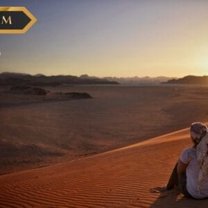 Una persona che indossa un foulard siede su una duna di sabbia al tramonto, affacciandosi su un vasto paesaggio desertico. Il cielo è limpido e la scena di pace sembra senza tempo. I loghi "PREMIUM", "sbs viaggi" e "Bozza automatica" appaiono negli angoli superiori.