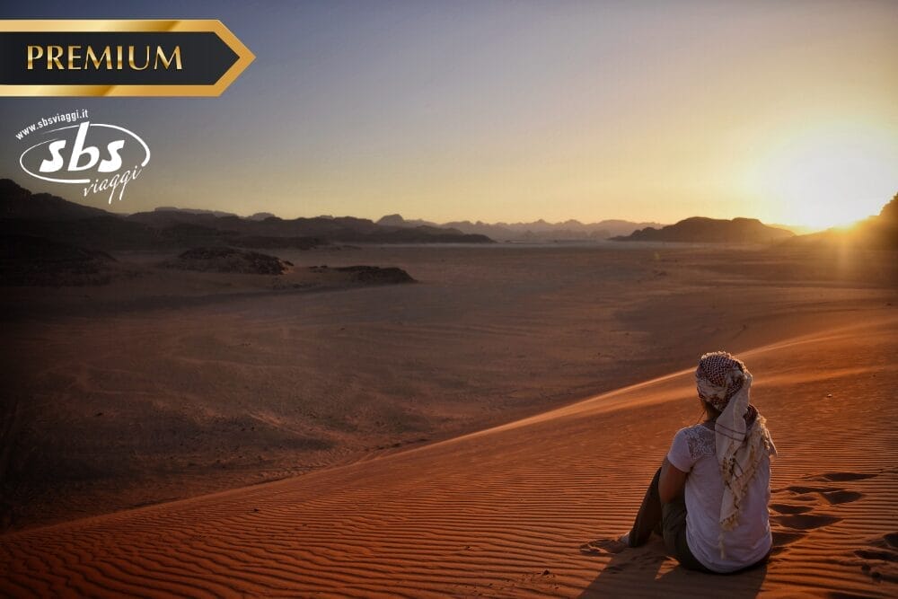 Una persona che indossa un foulard siede su una duna di sabbia al tramonto, affacciandosi su un vasto paesaggio desertico. Il cielo è limpido e la scena di pace sembra senza tempo. I loghi "PREMIUM", "sbs viaggi" e "Bozza automatica" appaiono negli angoli superiori.