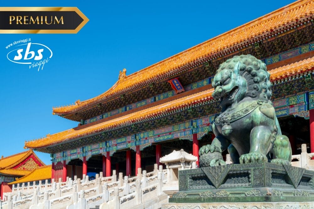 Una statua di bronzo di un leone si trova davanti a un edificio cinese tradizionale, ornato con un tetto dorato, sotto un cielo azzurro e limpido. Nell'angolo superiore compaiono la scritta "PREMIUM", il logo "SBS Viaggi" e "Bozza automatica".