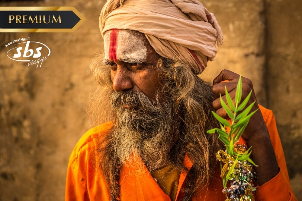 Un uomo anziano con capelli lunghi e barba, che indossa una tunica arancione e un turbante, tiene in mano una pianta frondosa. La sua fronte è segnata da vernice bianca e rossa. L'immagine presenta un'etichetta "PREMIUM", il logo "sbs viaggi" e un'etichetta Bozza automatica.