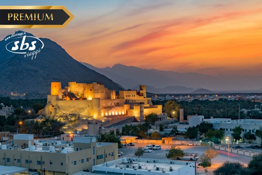 Veduta di un forte storico illuminato al tramonto con le montagne sullo sfondo e una città in primo piano. L'immagine include i loghi "PREMIUM", "sbs viaggi" e "Bozza automatica" nell'angolo in alto a sinistra.