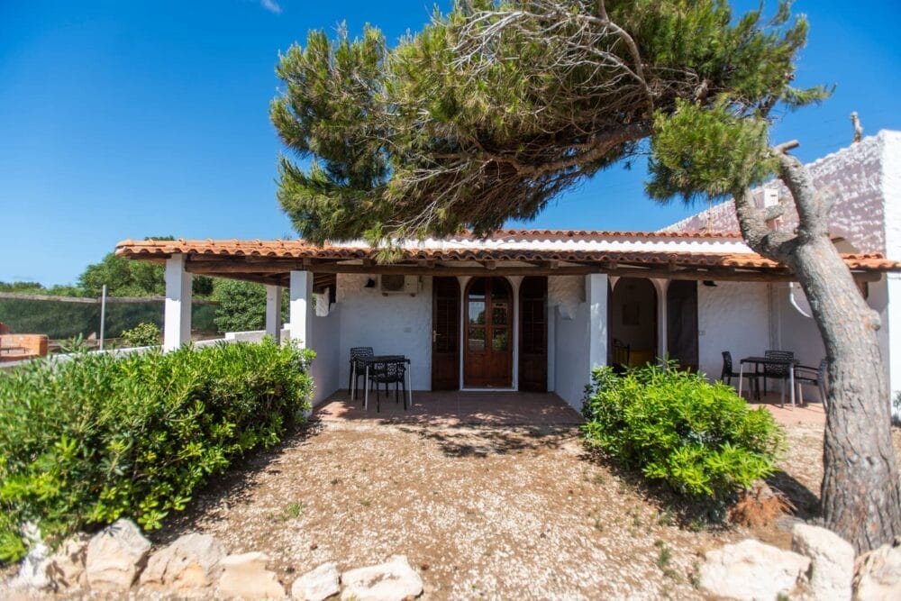 Una piccola casa bianca con tetto di tegole e porte ad arco in legno si trova sotto un cielo azzurro e limpido. Tavoli e sedie all'aperto invitano al relax, mentre il verde e un albero completano questo tranquillo ambiente di Bozza automatica.