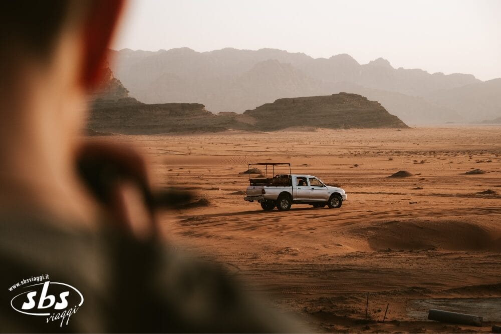 Un pick-up bianco si trova in una vasta pianura sabbiosa del deserto con montagne rocciose sullo sfondo. Il primo piano sfocato mostra una persona che guarda verso il camion, pronta per un tour con Tende di Stelle. Il logo di SBS Viaggi è visibile nell'angolo in basso a sinistra.