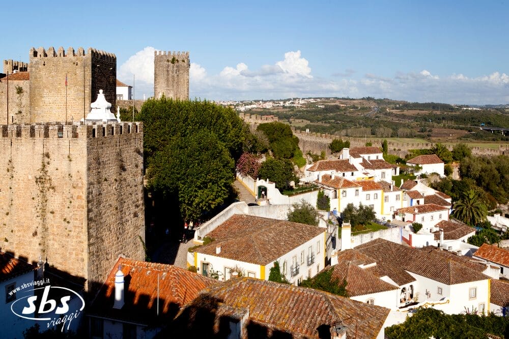 Una vista panoramica di Óbidos, in Portogallo, con il castello medievale in pietra, le case imbiancate a calce con i tetti rossi, le strade tortuose e la vegetazione lussureggiante sotto un cielo azzurro e limpido: una tappa incantevole del viaggio del Portogallo Express.